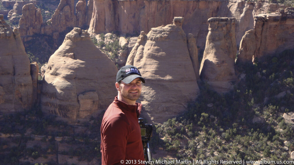 A man in a red fleece and dark cap stands before rounded red-rock canyon formations.