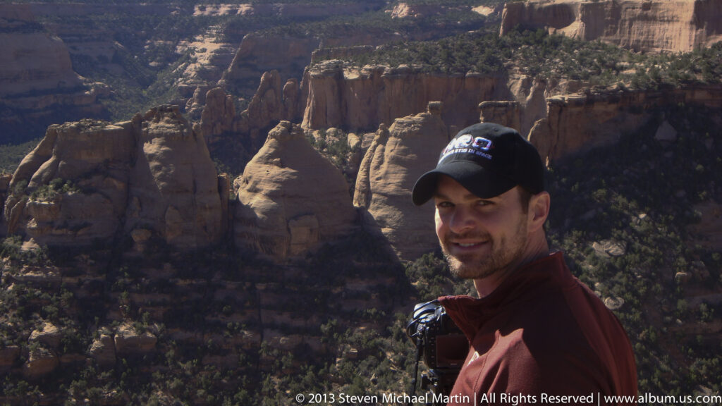 A close view of a man holding a camera, with red-rock canyon walls and shrubs behind him.