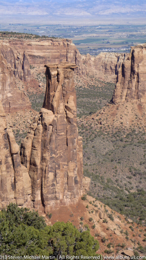 Tall red-rock towers rise from a broad canyon beneath a distant flat-topped plateau and blue sky.