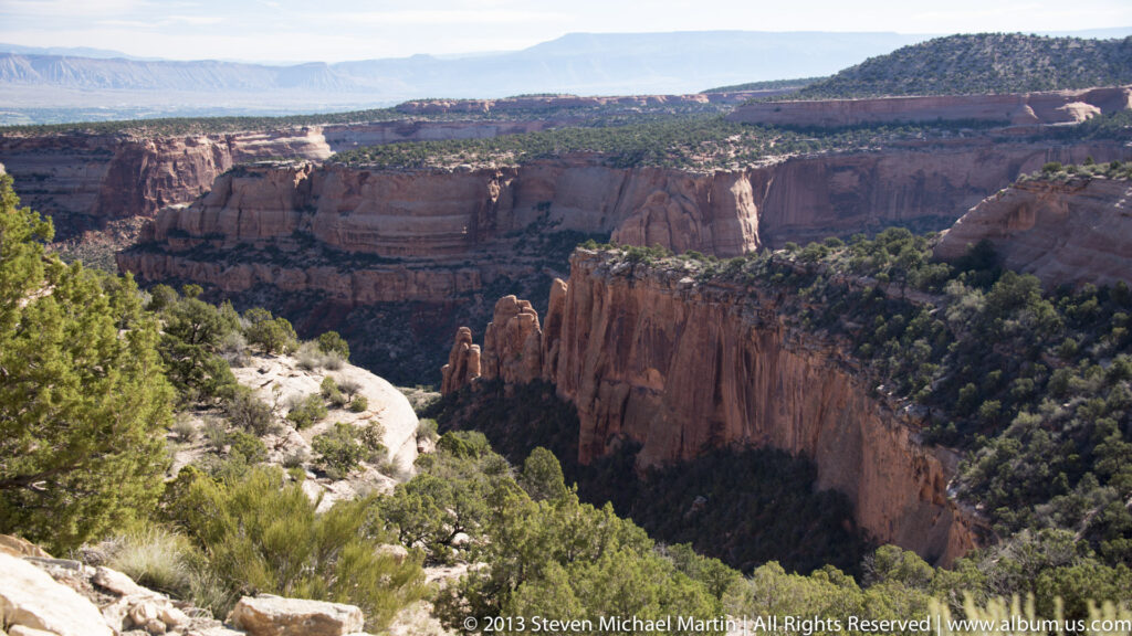 A rocky, shrub-lined rim overlooks a deep canyon with a projecting rock promontory and layered walls.