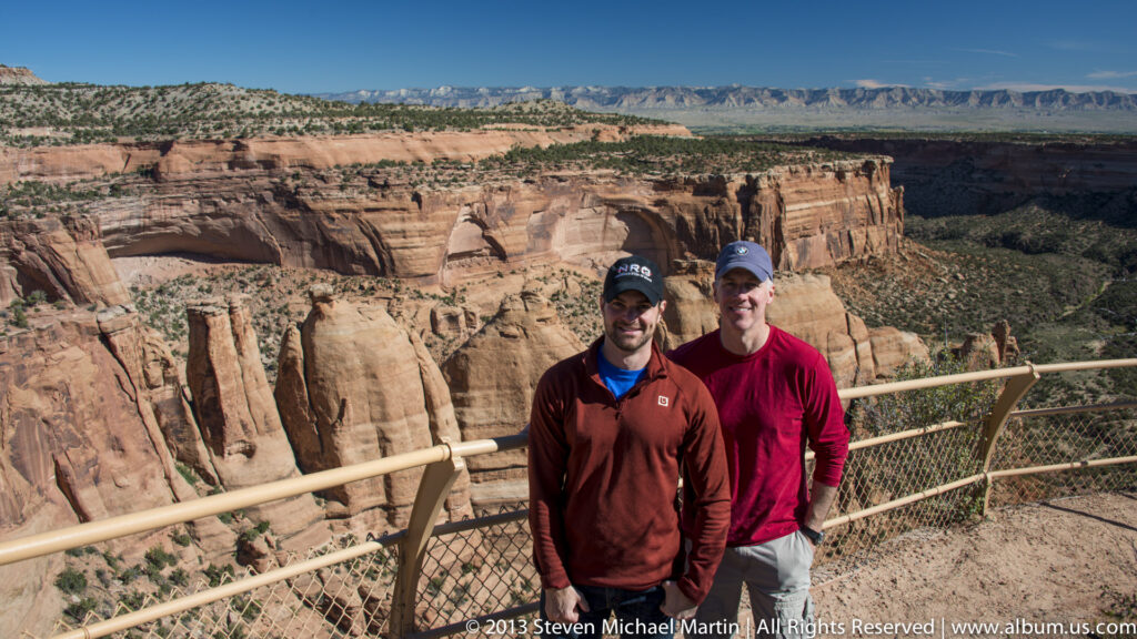 Two men pose behind a metal safety railing above a canyon of red-rock towers.
