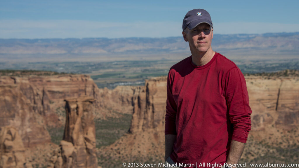 A man in a red shirt and dark cap stands before a softly focused canyon and distant plateau.