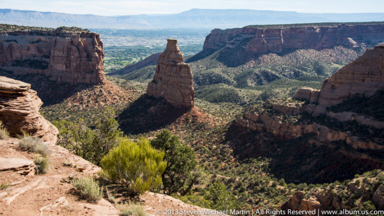 A narrow rock tower stands alone in a broad canyon surrounded by cliffs, shrubs, and distant mesas.
