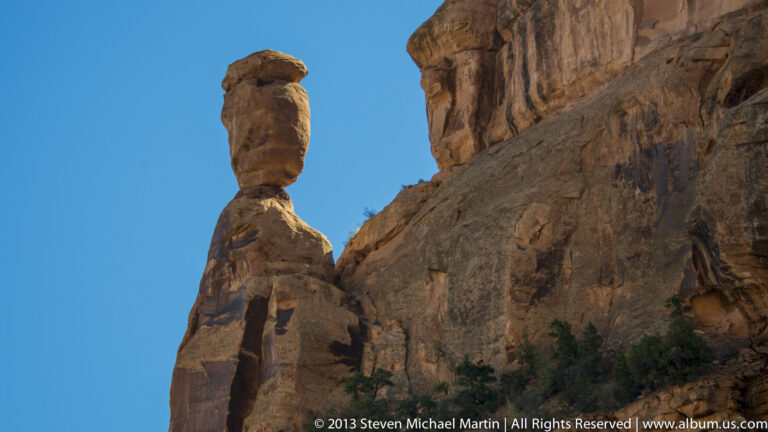 A rounded rock spire rises beside a massive cliff wall against a clear blue sky.