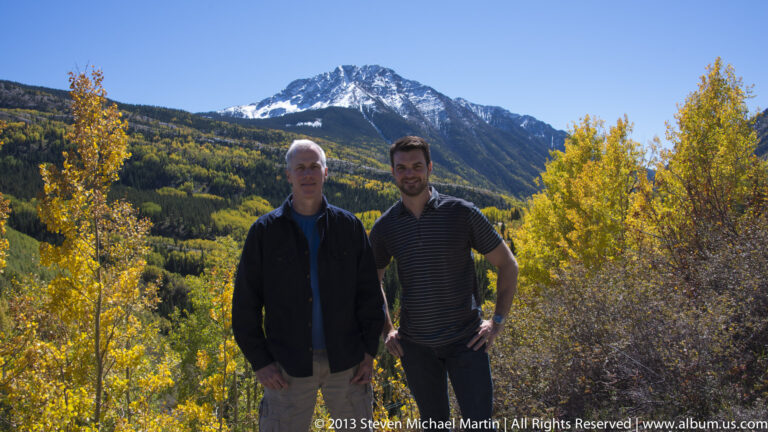 Two men pose among golden aspens with a snow-dusted peak behind them.