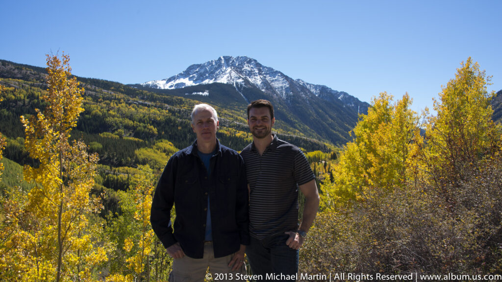 Two men stand closer to the camera, framed by yellow aspens and a snow-streaked mountain.
