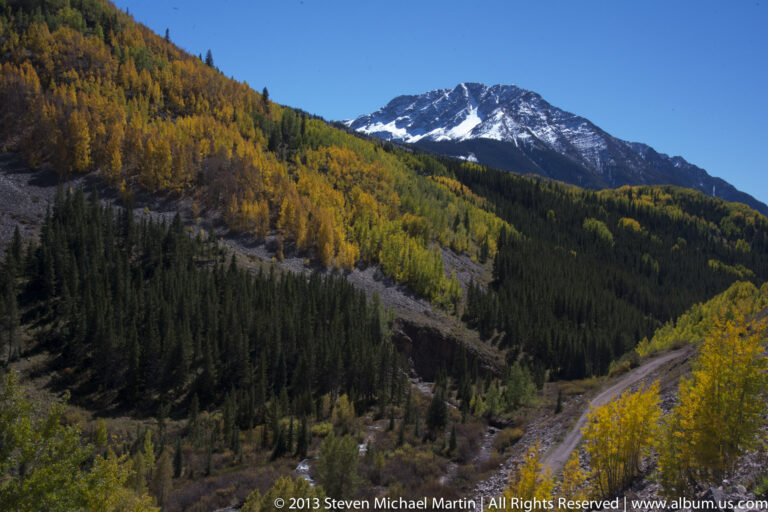 A curving road borders a forested slope of golden aspens beneath a snow-dusted peak.