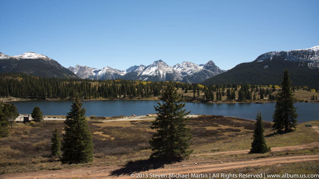 A mountain lake lies below snow-dusted peaks, with conifers along the shore and in the foreground.