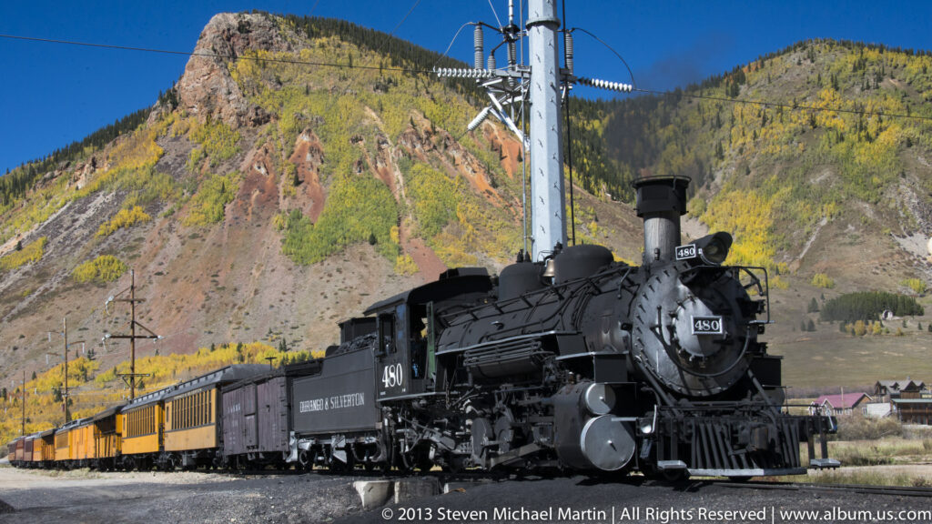 A black steam locomotive pulls yellow passenger cars below an autumn-colored mountain slope.