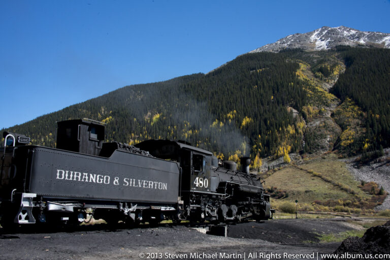 A black locomotive numbered 480, lettered “Durango & Silverton,” stands below a forested mountain.