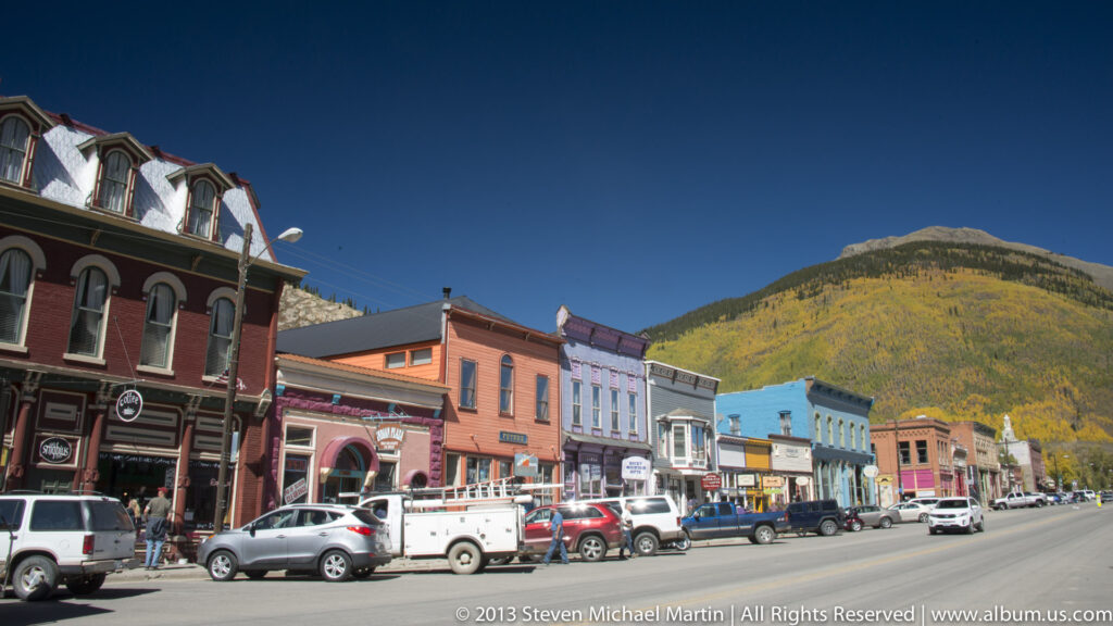 Colorful storefronts and parked cars line a town street below a golden mountain slope.