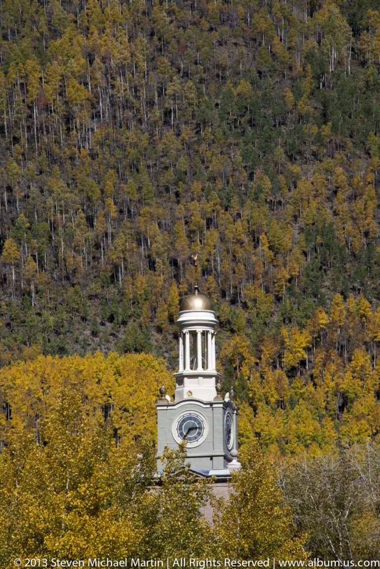 A white clock tower rises above golden trees in front of a wooded hillside.