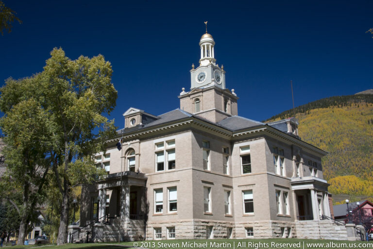 A pale civic building with a central clock tower stands among autumn trees below a wooded hill.