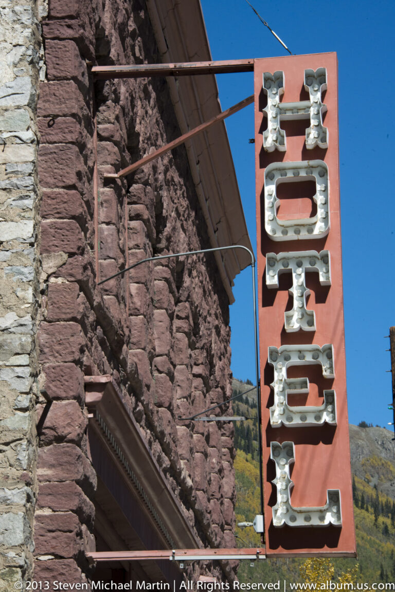 A vertical red sign with raised silver letters spelling HOTEL hangs beside a stone wall.
