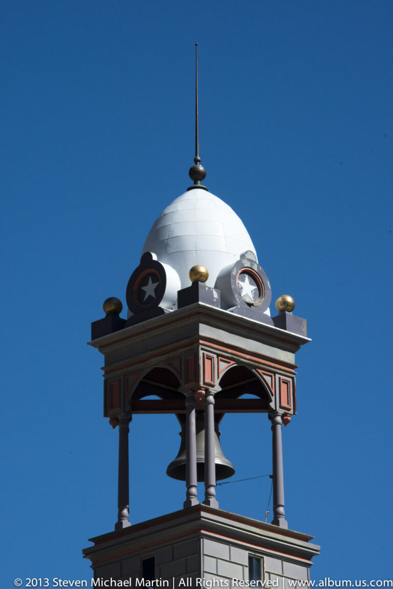 An open-sided cupola with a domed roof, star ornaments, and a hanging bell stands against blue sky.