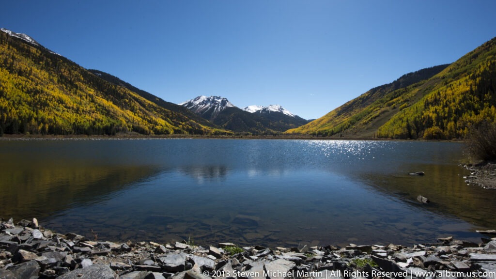 A lake stretches between forested autumn slopes toward snowy peaks at the end of a mountain valley.