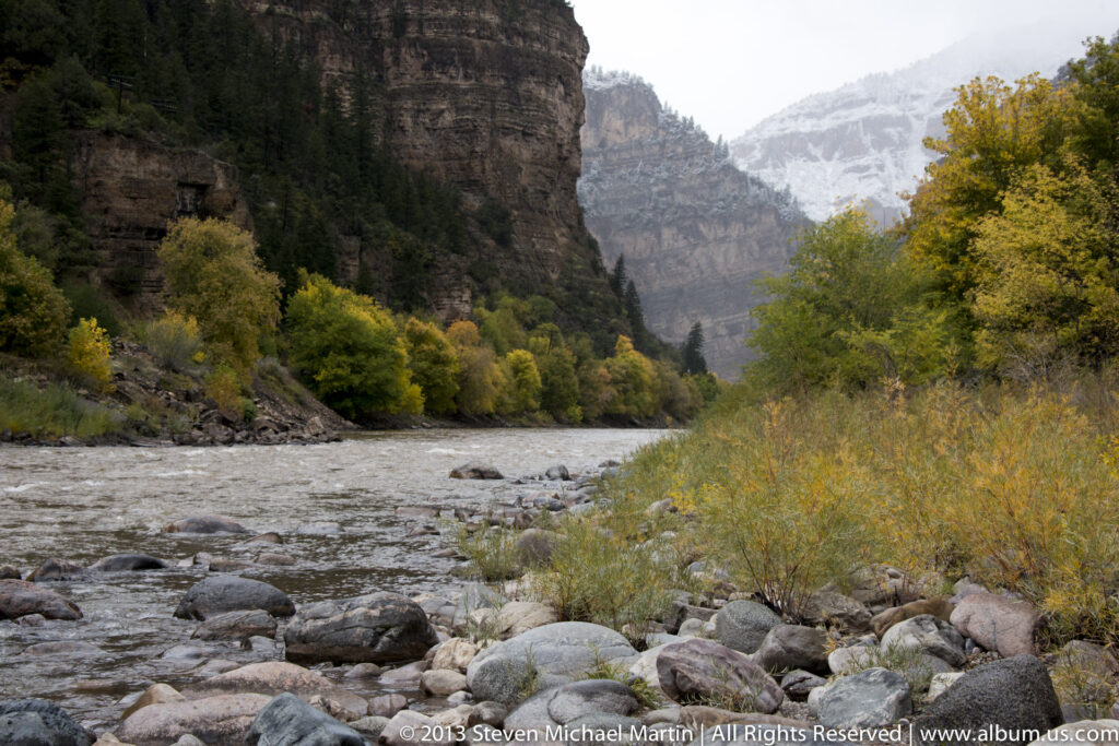 A river flows through a rocky gorge beneath dark cliffs, autumn shrubs, and snowy peaks.