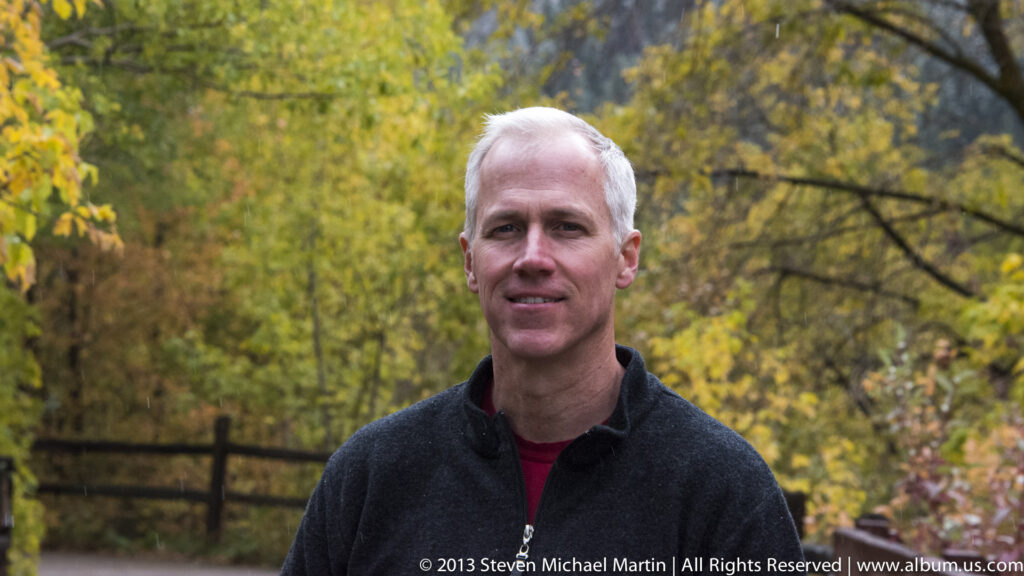 A man in a dark fleece stands before golden foliage and a wooden fence.