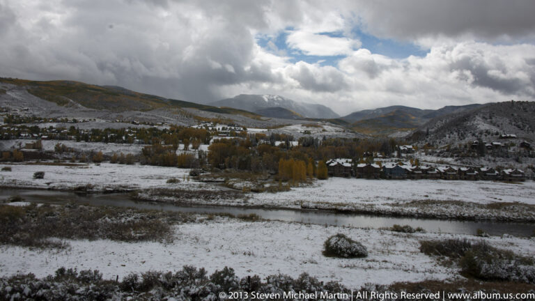 Fresh snow covers a valley with scattered buildings and autumn trees beneath clouded mountain ridges.