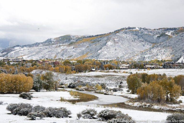 A shallow creek crosses a snowy meadow toward golden trees and buildings beneath a pale mountain slope.