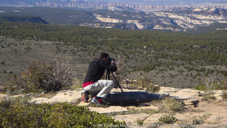 A kneeling visitor photographs distant layered canyon walls with a camera on a tripod.