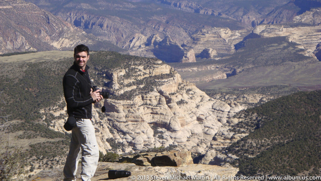 A man holding a camera stands above pale, layered canyon walls and dark scrub-covered slopes.