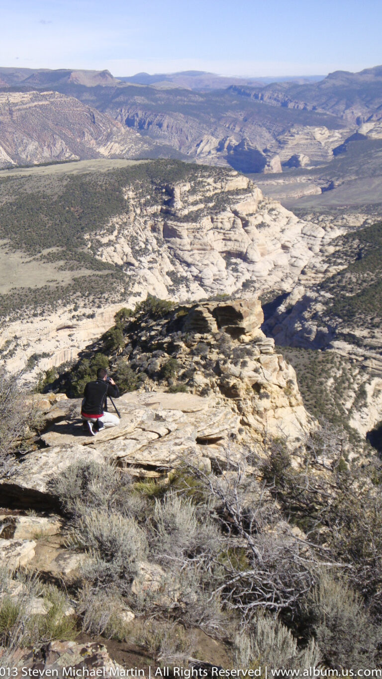 A small crouching visitor rests on a rocky rim above a broad, shrub-covered plateau and distant ridges.