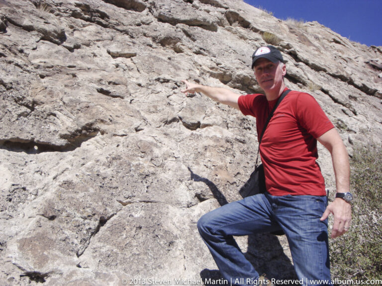 A seated man points toward a small mark on a pale, fractured rock face.