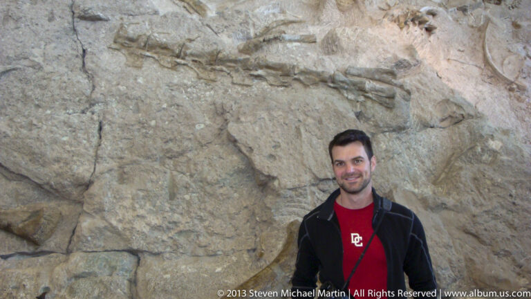 A man in a black jacket and red shirt stands before a pale, angular rock wall.