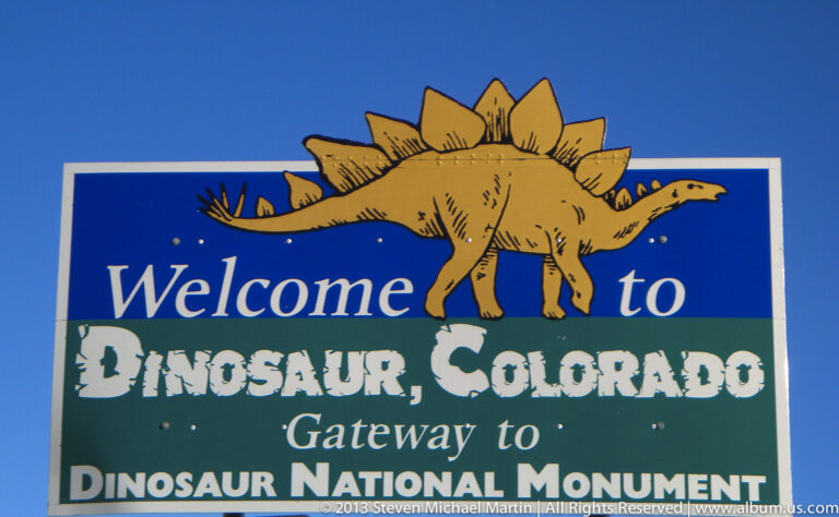 A blue and green welcome sign reads “Dinosaur, Colorado” and “Gateway to Dinosaur National Monument” beneath a stegosaurus.
