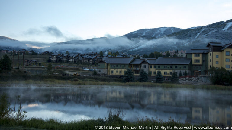 Multistory buildings reflect in a still pond as mist hangs along a mountain slope behind them.