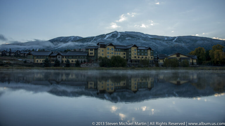 A broad row of buildings and mountain ridge reflects in a still pond beneath a pale blue sky.