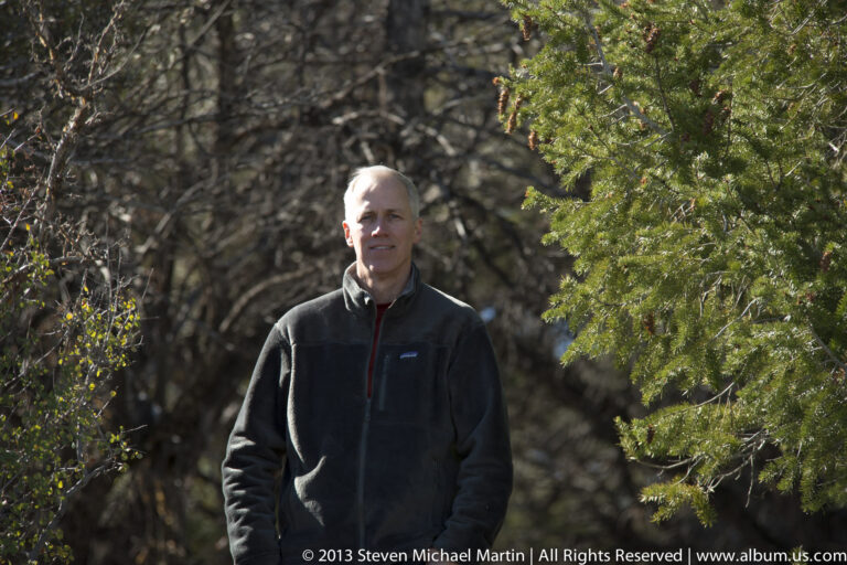A man in a dark fleece stands among tree trunks and dense branches in dappled light.