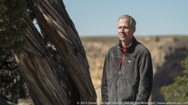 A man stands beside a large twisted trunk with dry scrubland visible behind him.