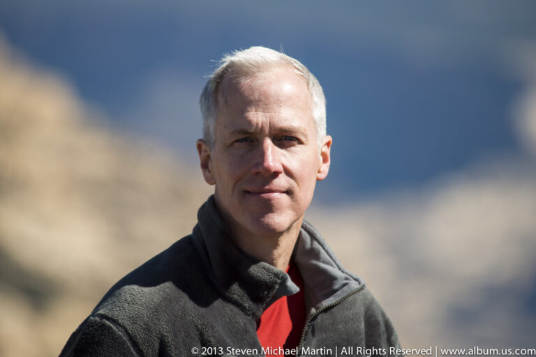 A close portrait of a man in a gray fleece against a softly blurred outdoor background.
