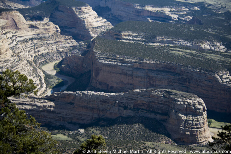 A green-blue river curves beneath a high layered cliff and scrub-covered canyon benches.