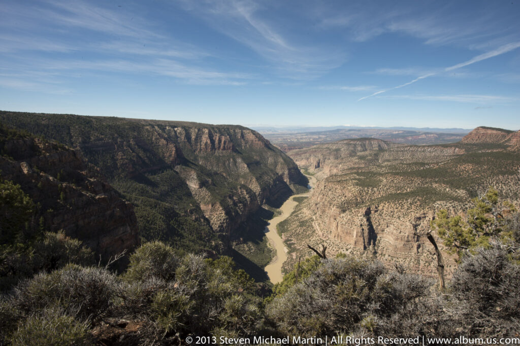 A winding river divides shadowed and sunlit canyon walls beneath a blue, cloud-streaked sky.