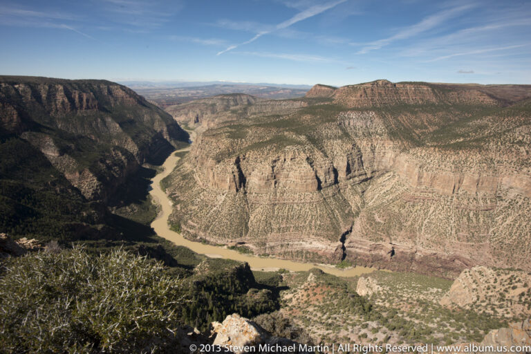 A river curves below a sunlit, gullied cliff, with shrubs and small trees in the foreground.