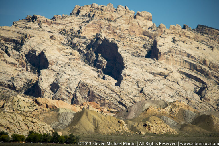 Pale sandstone cliffs with a dark recess rise above ocher slopes and a line of trees.