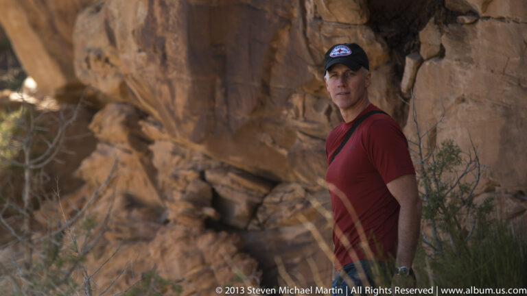 Man in a red shirt and cap stands beside a tall sandstone wall inside a shaded alcove.