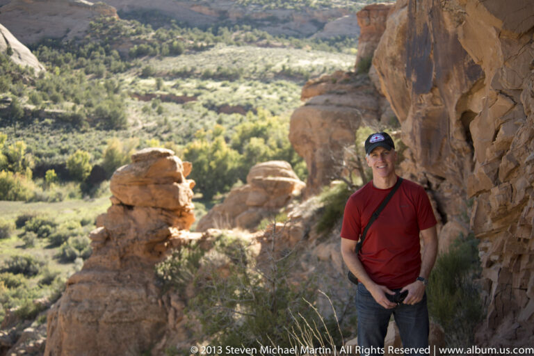 Man in red stands beside sandstone pillars, with a green valley visible through the opening.