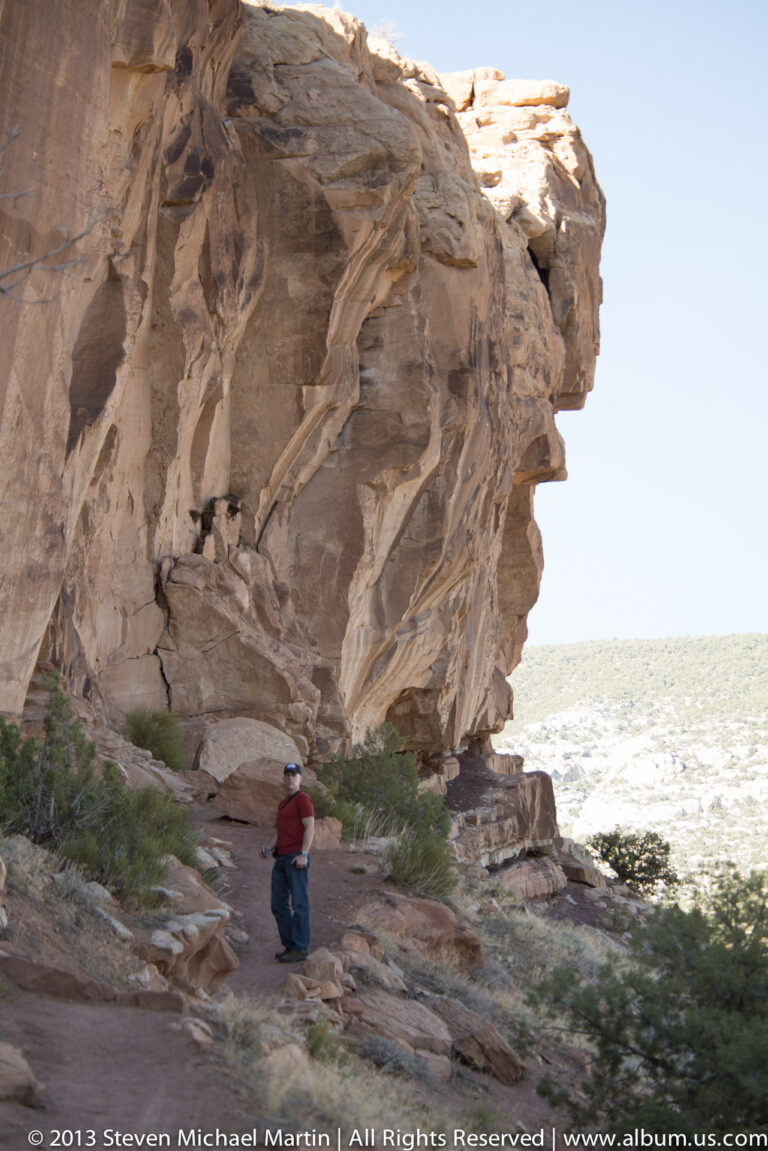Small visitor stands beneath a huge sandstone overhang beside a sunlit open plain.