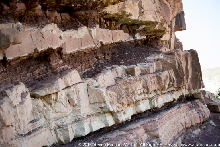 Cream, red, and gray sandstone layers form a fractured ledge with a dark recess below.