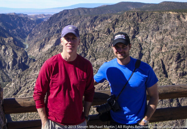 Two men in red and blue shirts pose beside a wooden rail with the canyon walls behind them.