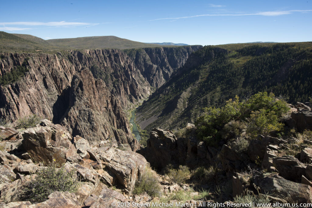 Rocks and shrubs frame a down-canyon view toward a narrow river between sunlit and shadowed cliffs.