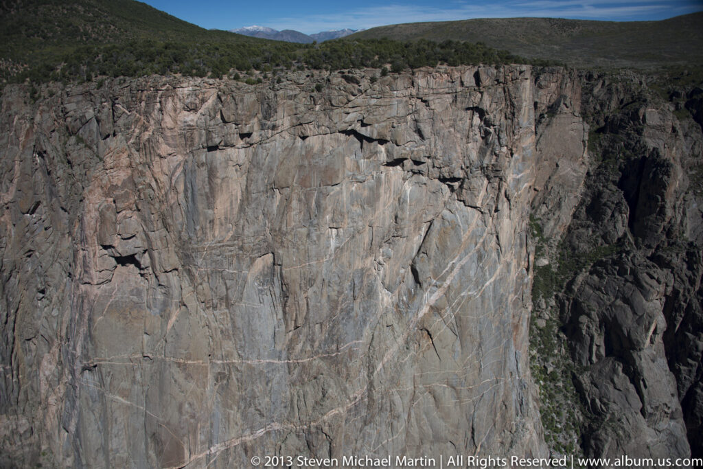 A steep fractured cliff fills the view, with vertical cracks and ledges beneath a narrow strip of rim vegetation.