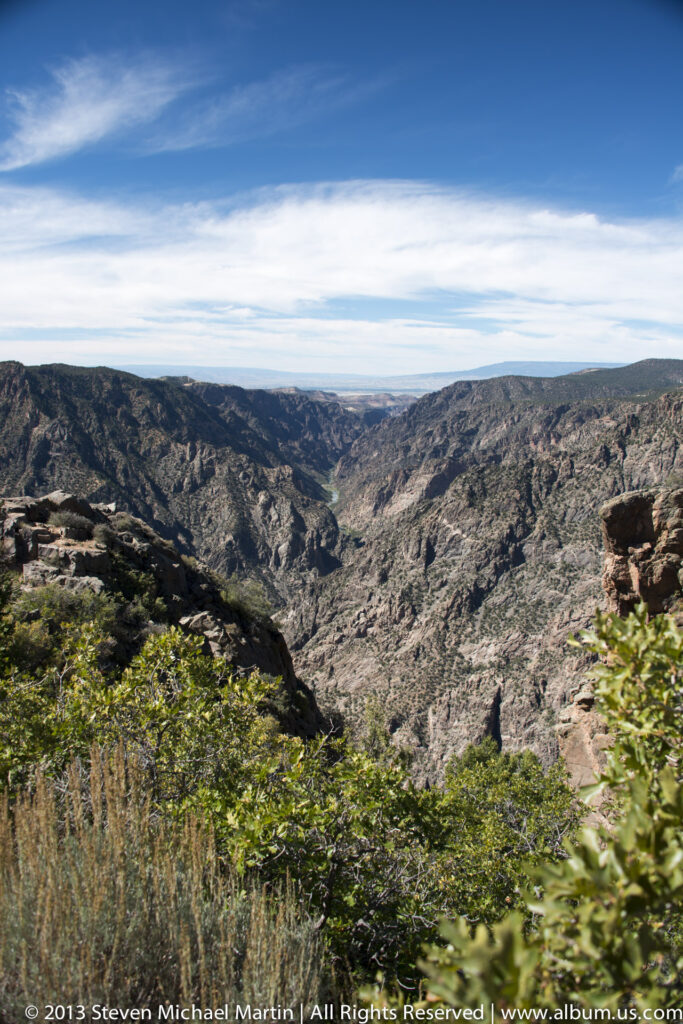 A winding canyon narrows toward a distant river, framed by sunlit shrubs and rugged rock in the foreground.