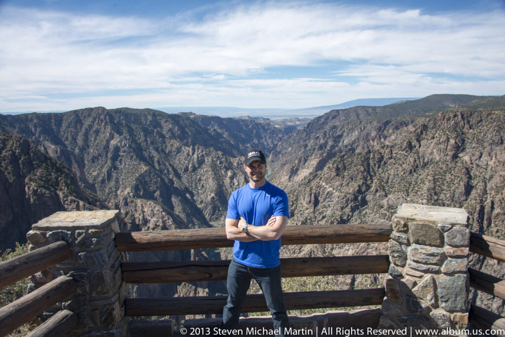A man stands behind a timber rail between stone pillars, with the canyon and distant plateau beyond.