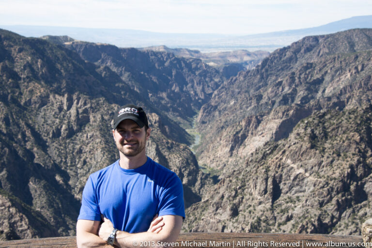A man in a blue shirt stands with folded arms at a wooden rail above the deep canyon and distant river.