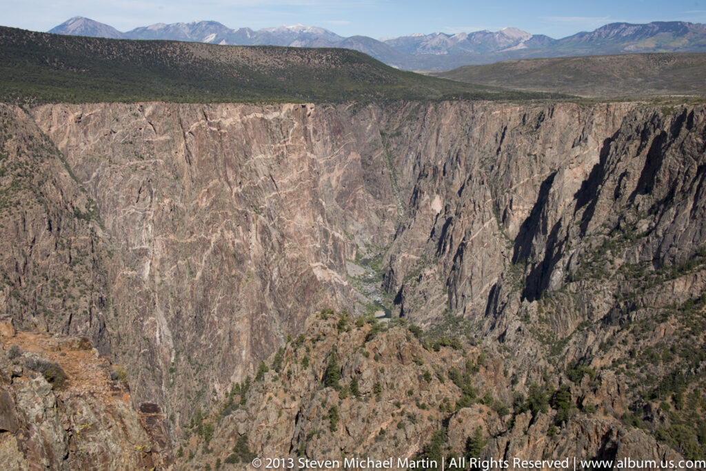 A wide canyon vista reveals layered cliffs, a distant ridge, and sparse shrubs along the rocky foreground.